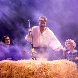 A man with a gardening fork spears a hay bail. There is a purple haze in the air. Two women look on, behind. 