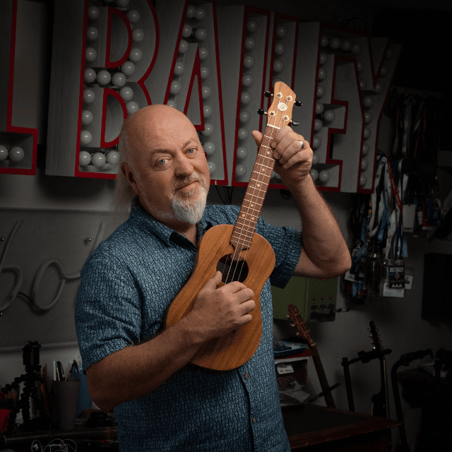Bill Bailey holding a ukelele upright and looking at the camera.