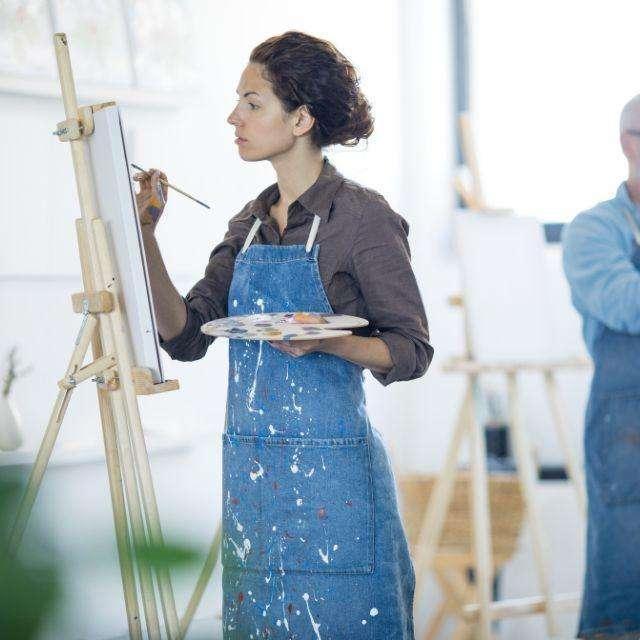 Someone painting on an easel wearing a denim apron splattered in paint.