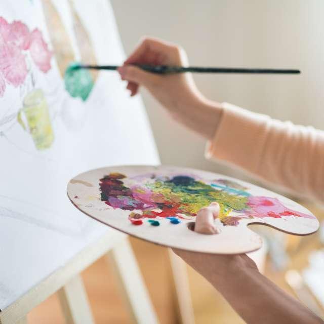 Someone painting on an easel holding a wooden paint palette with many different colours on it.
