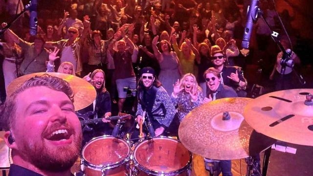 A drummer in a band is taking a selfie on stage showcasing the other band members and the audience.