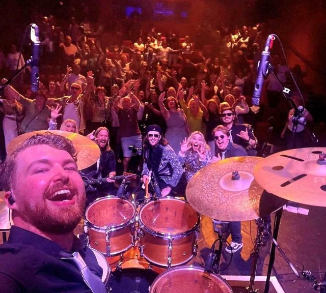 A drummer in a band is taking a selfie on stage showcasing the other band members and the audience.