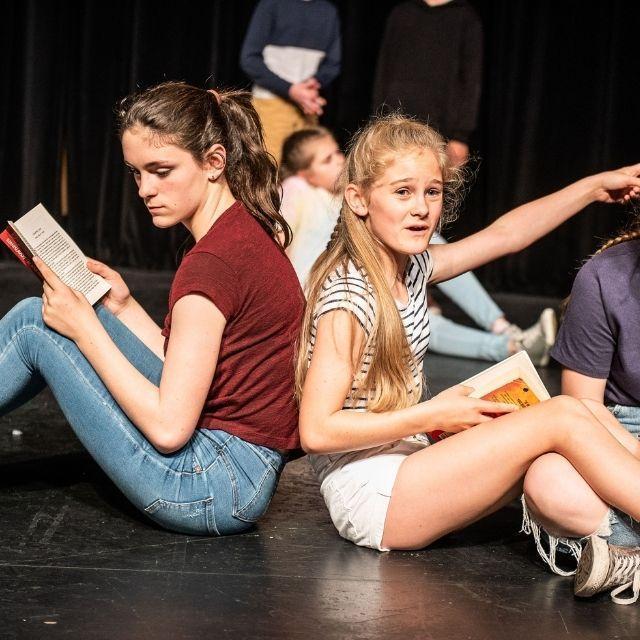 A group of young people doing a performance. They are sat on stage back to back reading books.