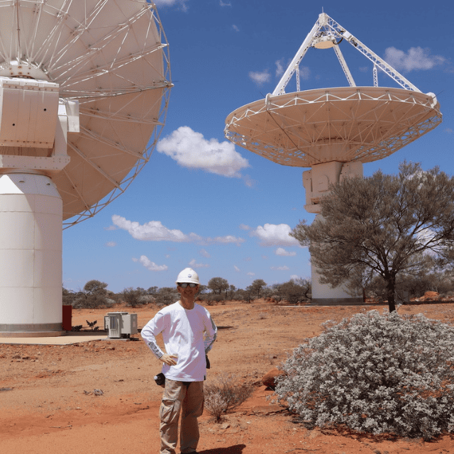 A person wearing protective clothing, including a white hard hat, sunglasses and cargo pants standing outside in a desert-like environment beneath two giant white satellite dishes.