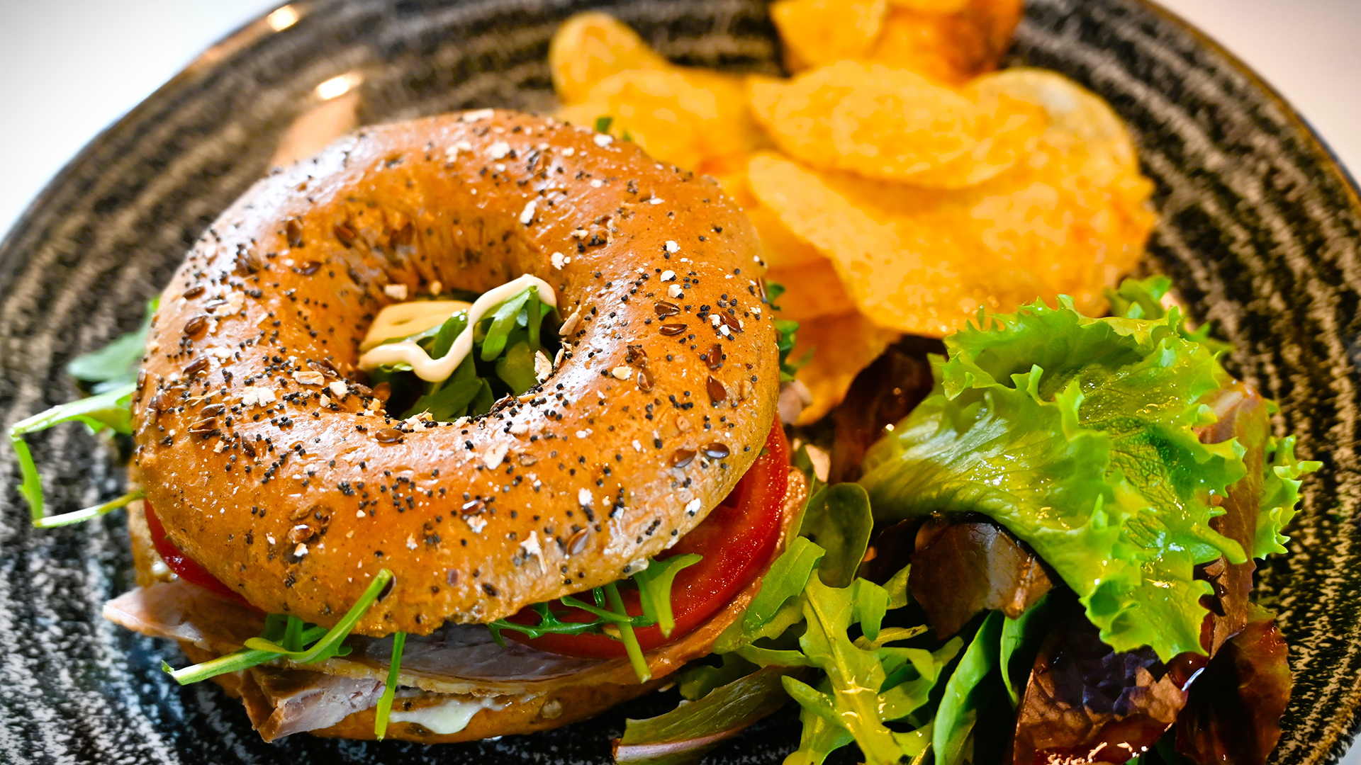 A close up of a seeded BLT bagel with salad and crisps on the side.